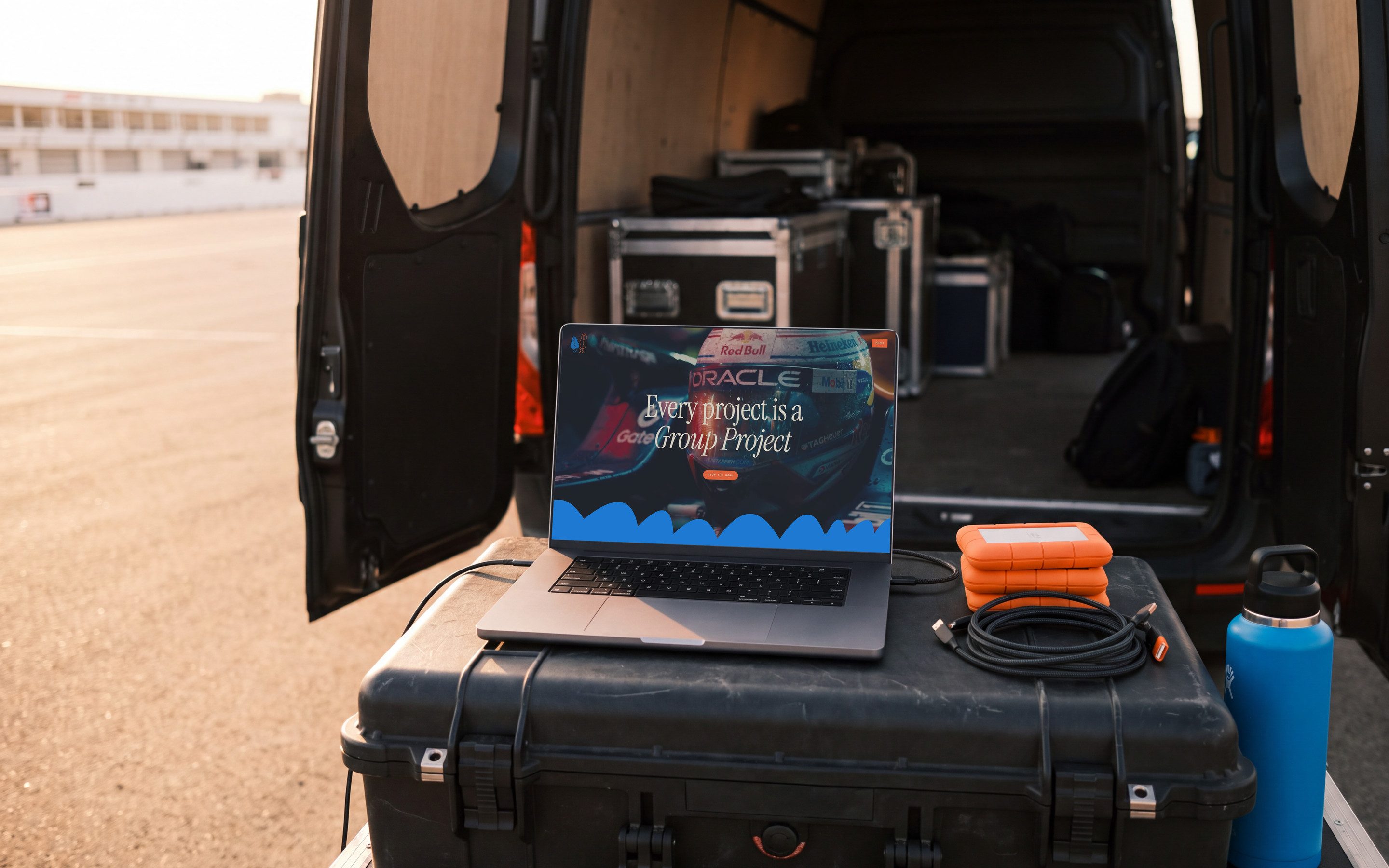 The Group Project homepage open on a laptop, set on a hard case at the tailgate of a production van, a race paddock behind it in late sun.