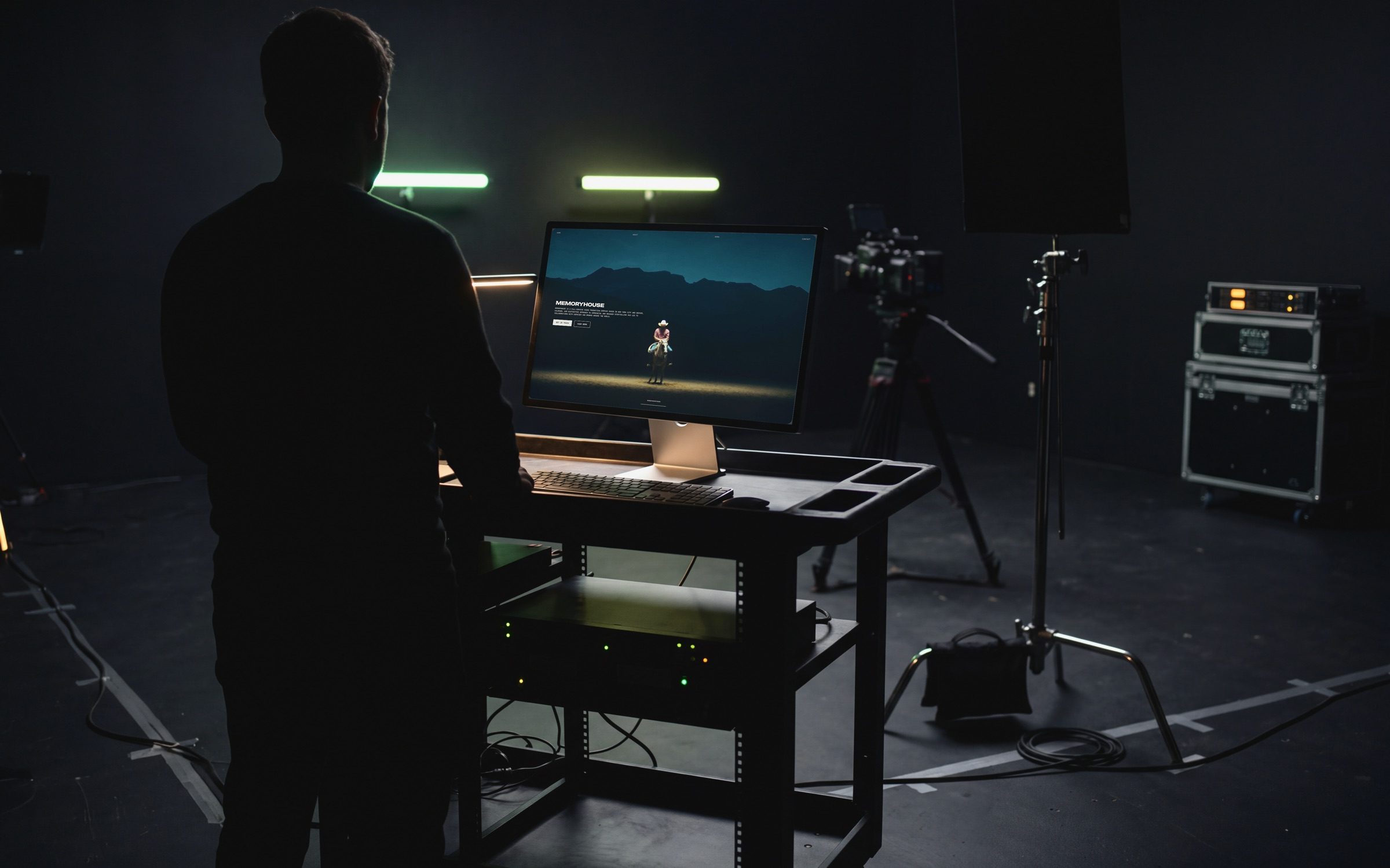 The Memory House homepage on a display in a dark sound stage, a rider lit alone in a pool of light, a figure watching from behind beside a camera on its tripod.