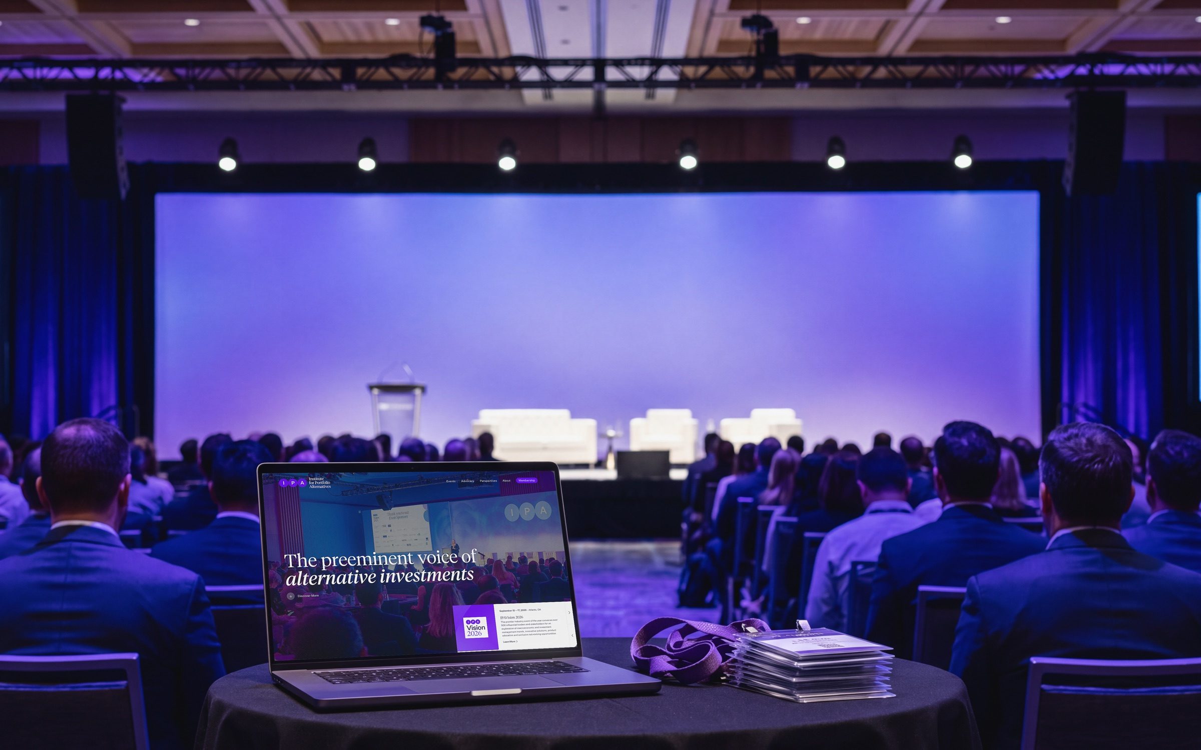 The IPA homepage open on a laptop at a front table in a conference ballroom, the audience seen from behind facing a stage washed in violet light.