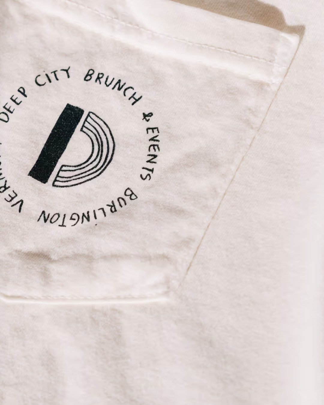 A cream pocket T-shirt printed with a round stamp that reads Deep City Brunch & Events, Burlington, Vermont, cut close from the Deep City home page.